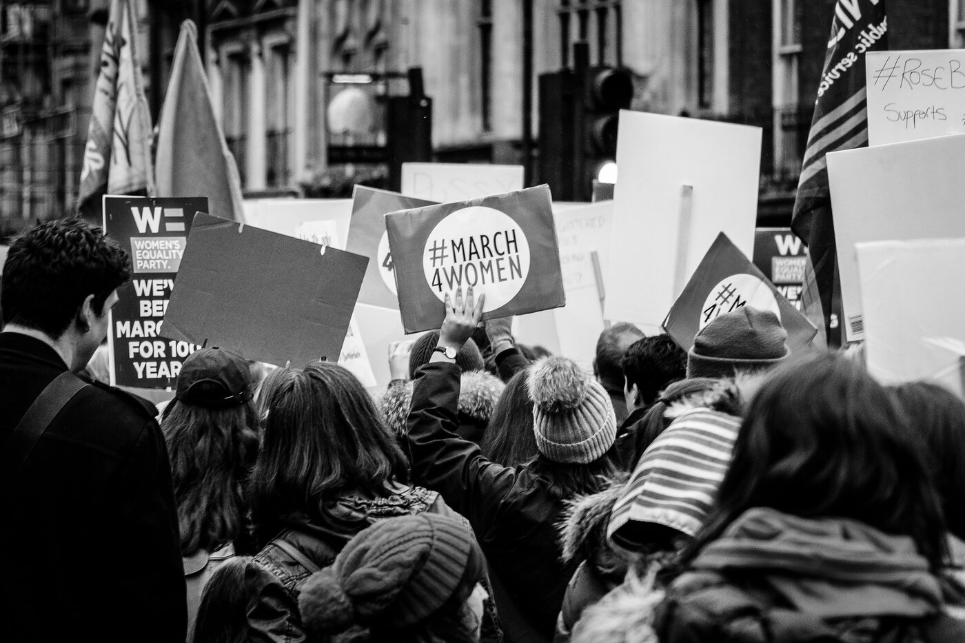 Black and white image of of a group of protesters, seen from behind. All are holding up placards, the most visible one in the cenbtre of the image reads 'March 4 Women'